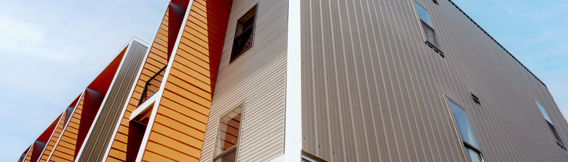 Low-angle view of a modern multi-story building with orange wood-style siding, white trim, and tan vertical metal panels, set against a blue sky with thin clouds.