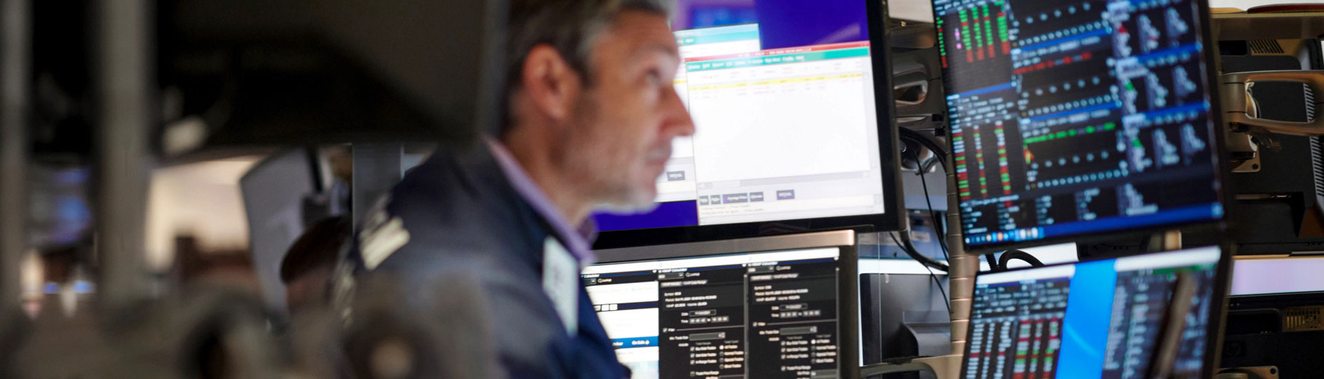 A trader on a stock exchange floor surrounded by multiple monitors displaying live financial data, charts, and trading information.