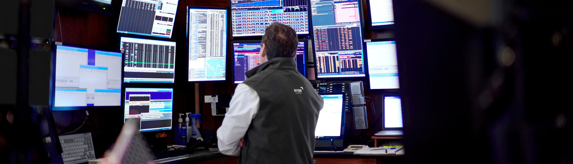A trader on a stock exchange floor surrounded by multiple monitors displaying live financial data, charts, and trading information.